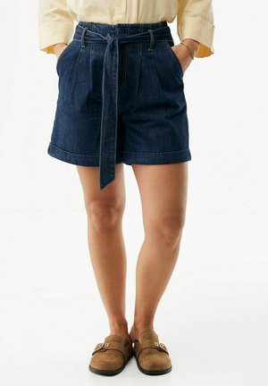 Kobieta ubrana w ciemne szorty jeansowe z paskiem, jasnożółtą koszulę włożoną do środka i brązowe wsuwane buty, stojąca z rękami w kieszeniach.