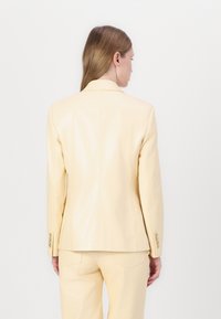 Blazer in pelle giallo chiaro con una forma sartoriale, apertura posteriore singola e dettagli ai polsini con tre bottoni. Texture liscia, design minimalista.