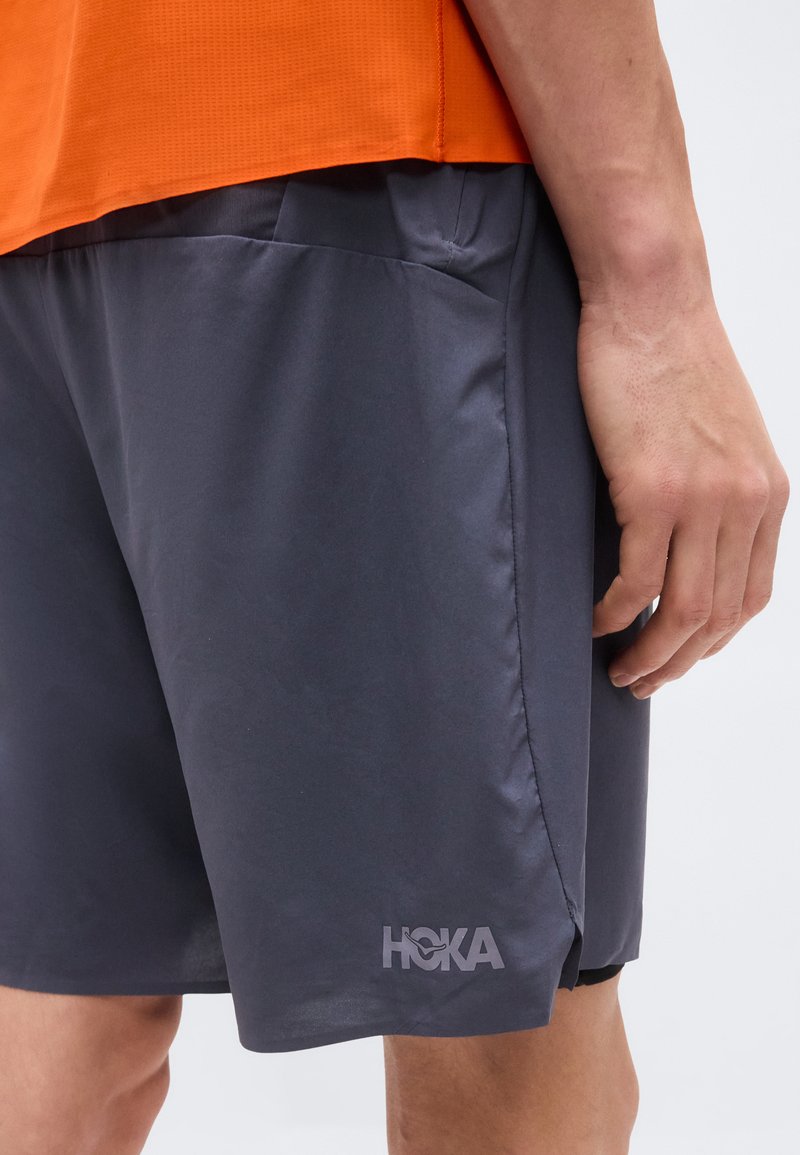 Nahaufnahme einer Person, die dunkelgraue HOKA-Shorts und ein oranges Shirt trägt, mit sichtbarer Seitentasche und Unterarm.