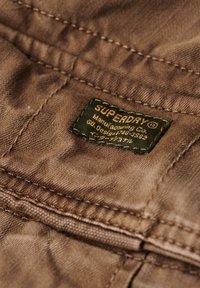 Bruine stof met een gestructureerd oppervlak. Bevat een groene label die erop is genaaid, waarop "SUPERDAY" en "Manufacturing Co." in contrasterende tekst staat.