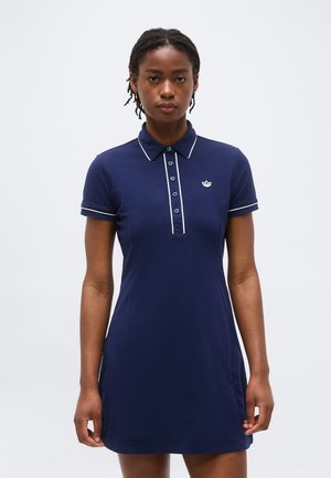 Tânără purtând o rochie polo bleu închis cu margini și nasturi albi, stând pe un fundal simplu, deschis la culoare.