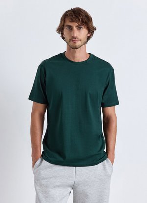 Uomo con capelli castani corti che indossa una maglietta verde scuro e pantaloni grigio chiaro, in piedi con le mani nelle tasche davanti a uno sfondo semplice.