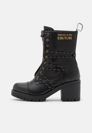 Czarny skórzany but do połowy łydki na obcasie blokowym, z wytrzymałą podeszwą, sznurowaniem z przodu oraz złotym logo "Versace Jeans Couture" po zewnętrznej stronie.