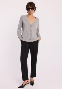Szary sweter z dzianiny z dekoltem w serek i sześcioma guzikami, zestawiony z czarnymi spódnicami plisowanymi i czarnymi szpilkami. Model stoi z rękami w kieszeniach.