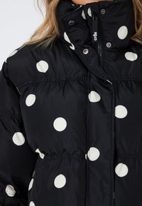 Svart polstret jakke med store hvite prikkete mønstre. Har høy krage, glidelås lukking og elastisk midje for en tettsittende look.