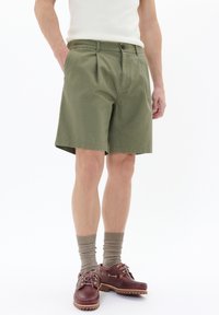 Shorts plisate de culoare verde măsliniu cu închidere cu nasture, fabricate dintr-un material ușor. Purtați cu pantofi bărcă cu talpă de cauciuc de culoare maro și șosete dublate bej.