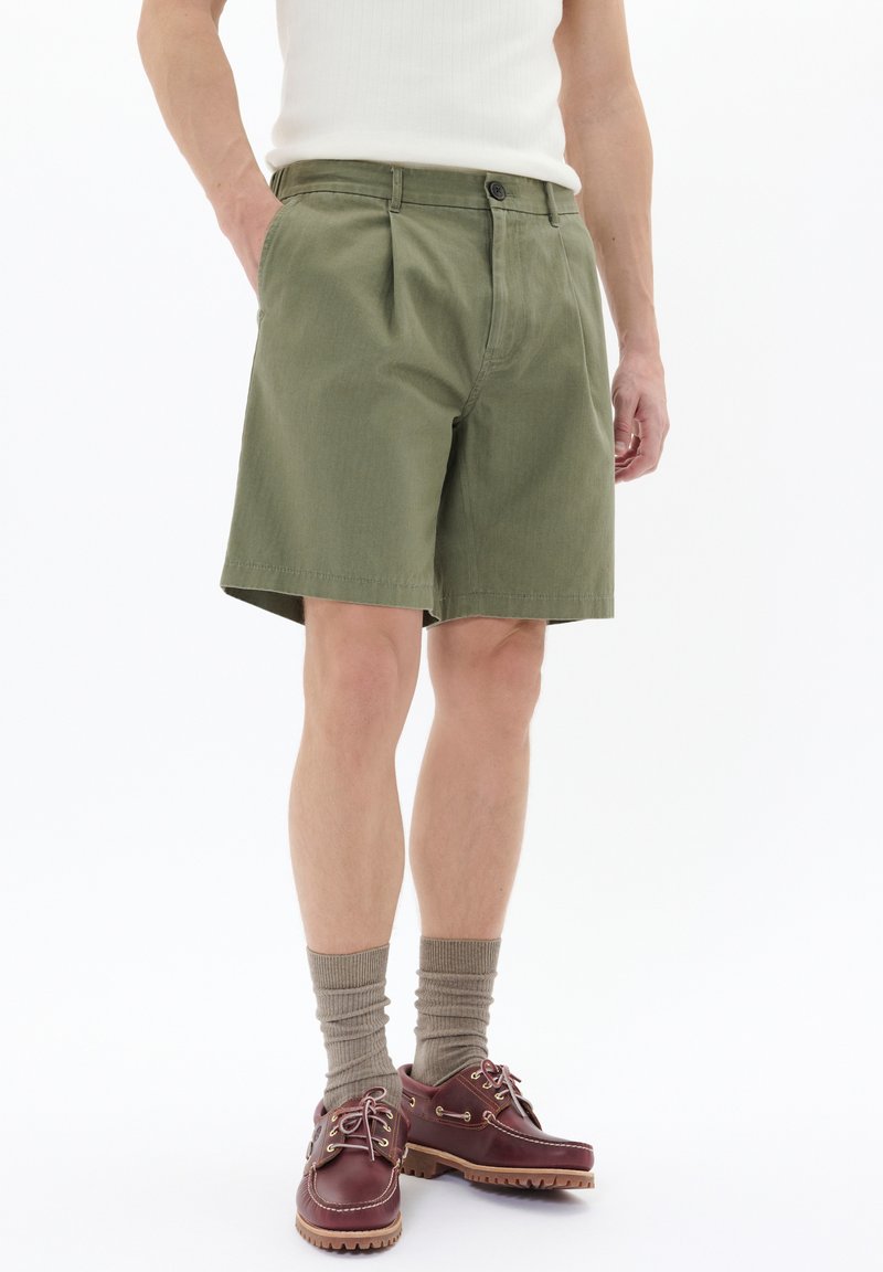 Shorts plisate de culoare verde măsliniu cu închidere cu nasture, fabricate dintr-un material ușor. Purtați cu pantofi bărcă cu talpă de cauciuc de culoare maro și șosete dublate bej.