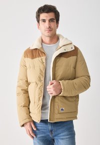 Chaqueta acolchada color beige con parte superior de pana, cuello de borreguito, cierre frontal y parche con el logo. Llevada sobre una camisa gris, combinada con jeans azules.