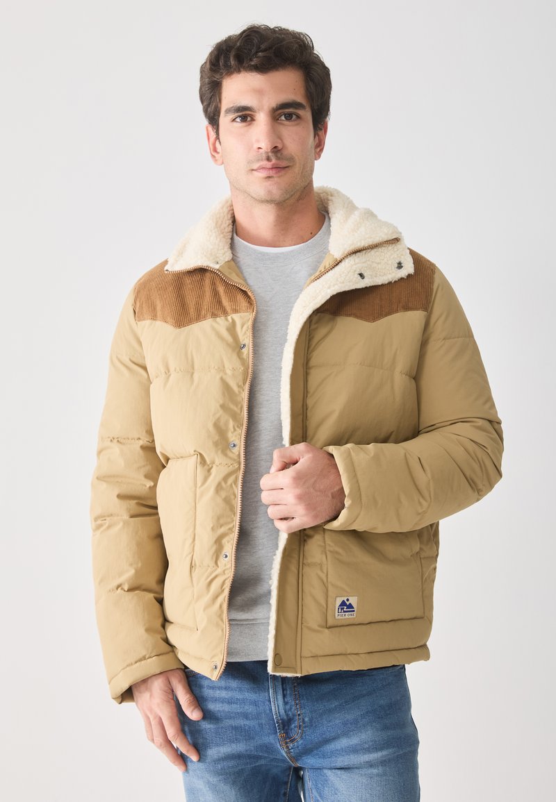 Beige pufferjack met een corduroy bovenstuk, schapenvacht kraag, ritssluiting aan de voorkant en een logo-embleem. Draag het over een grijze shirt, gecombineerd met blauwe spijkerbroeken.