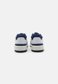 Fehér és sötétkék sportcipő sima bőr felső résszel, hálós részletekkel és textúrázott gumitalppal. Lacoste logó a saroknál.