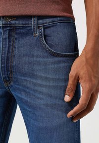 Ciemnoniebieskie dżinsy posiadają widoczne szwy, kieszeń z przodu oraz zapięcie na guzik w talii. Tekstura jest gładka i lekko wyblakła.