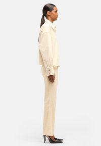 Camicia di cotone beige con maniche oversize e bottoni, abbinata a pantaloni beige aderenti e tacchi neri a punta, mostrata di profilo.