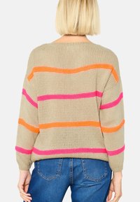 Beige gebreide trui met gedurfde horizontale strepen in roze en oranje, met een losse pasvorm en geribbelde manchetten. Gecombineerd met blauwe spijkerbroek.