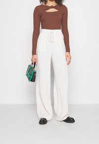 Kvinna klädd i en brun långärmad topp med u-ringning, lösa vita byxor med dragsko, svarta klumpiga skor, håller en svart handväska med gröna detaljer.