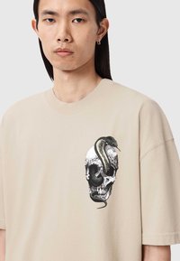 Camiseta beige de gran tamaño con un gráfico detallado de una calavera con una serpiente enrollada alrededor en colores negro y verde.