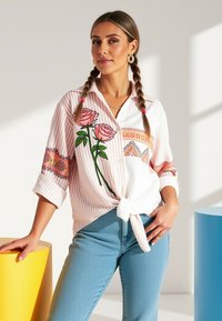 Biała i różowa koszula z guzikami w paski, ozdobiona haftem kwiatowym i z zawiązanym przodem. W połączeniu z jasnoniebieskimi dżinsami.