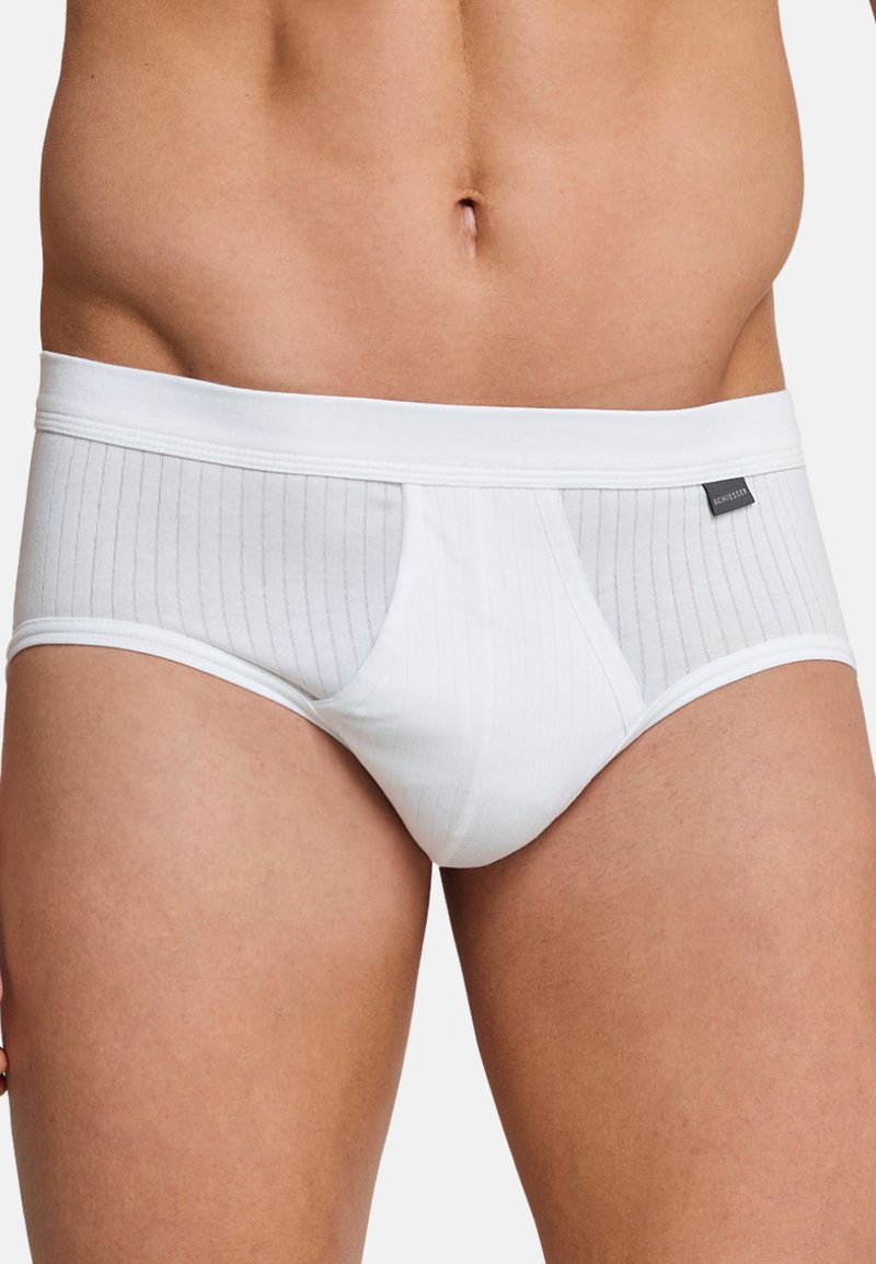 Torso masculin portant un slip blanc côtelé avec une étiquette de marque noire sur la hanche droite, debout devant un fond uni.