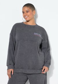 Grijze oversized sweatshirt met ribgebreide manchetten en zoom. "Studio Untold" en "Travel Basics" geborduurd in contrasterende kleuren op de borst.
