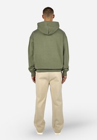 Felpa verde con una vestibilità comoda, tasca a canguro e cappuccio regolabile, abbinata a pantaloni beige a gamba larga e sneakers bianche.