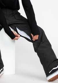 Pantalons de neige noirs avec des aérations zippées, doublure en maille et logo subtil. Porté avec un haut à manches longues et des chaussures noires avec des accents blancs.