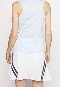 Person som bär en ljusblå ärmlös topp och en vit kjol med svarta diagonala ränder, sedd bakifrån mot en enfärgad bakgrund.