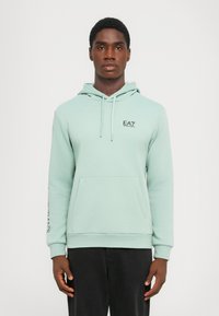 Mint green hoodie with front pocket, drawstrings, and black EA7 logo on chest. Long sleeves feature a printed design. Soft fabric texture.