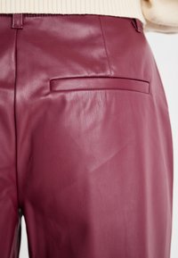 Pantalon en cuir bourgogne avec une texture lisse, une coupe ajustée et une poche arrière unique sans matériel visible ni détails de couture.