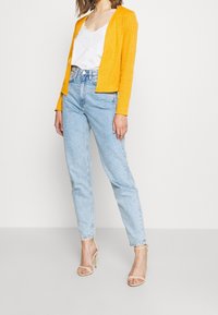 Světlý žlutý žebrovaný cardigan se rovným střihem, kombinovaný s světle modrými džíny s vysokým pasem a nahými sandály na vysokém podpatku. Pod ním je bílý top s ramínky.