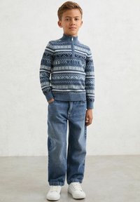 Niepodrabialny niebieski sweter z białymi geometrycznymi wzorami i wysokim kołnierzem, zestawiony z wyblakłymi niebieskimi dżinsami i białymi sneakersami.