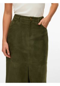 Femme portant une jupe en daim vert olive avec fente avant, bouton avant, passants de ceinture et la main dans la poche droite, associée à un haut blanc.