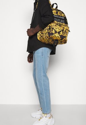 Sac à dos noir avec motif baroque doré, arborant un logo bien visible et des fermetures éclair, assorti à un jean bleu clair et des baskets blanches.
