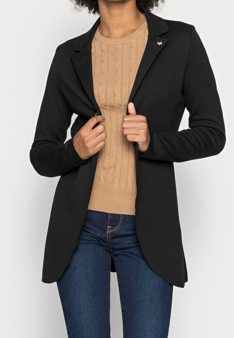Czarna, dopasowana marynarka o gładkiej fakturze, z zapięciem na jeden guzik i małym ozdobnym akcentem, nałożona na jasnobrązową dzianinową bluzkę.