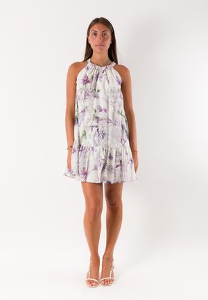 Donna che indossa un vestito bianco senza maniche con stampa floreale viola e sandali bianchi con tacco alto, in piedi davanti a uno sfondo bianco uniforme.