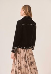 Mujer vistiendo una chaqueta negra bordada con detalles florales en los puños y el dobladillo, combinada con una falda beige con pliegues y estampado floral negro.