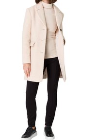 Manteau texturé rose clair avec un col cranté, trois boutons à l'avant et deux poches, associé à un haut beige et un legging noir.