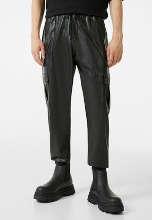 Hombre con pantalones cargo negros de piel sintética con puños elásticos y botas negras gruesas hasta el tobillo, de pie frente a un fondo liso.
