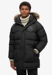 Chaqueta de pluma negra con capucha con ribete de piel, que presenta un parche redondo de logo, cierre frontal a presión y dos grandes bolsillos. Diseño texturizado e insulated.