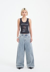 Czarny top na ramiączkach z graficznym wzorem, w zestawieniu z szerokimi jasnoniebieskimi dżinsami, prezentowany na modelce na białym tle.