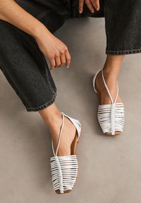 Sandales blanches à bout ouvert avec un design de bride tissée, semelle plate et détails minimalistes, portées avec un jean foncé sur une surface neutre.