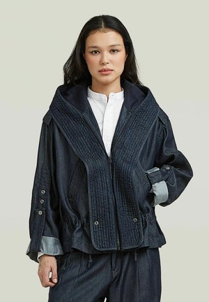Vrouw met donker haar draagt een oversized donkerblauwe doorgestikte jas over een wit overhemd met knopen op een lichte achtergrond.