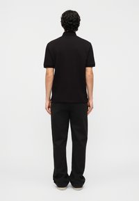 Άνδρας με σγουρά σκουρόχρωμα μαλλιά, που στέκεται πλάτη, φοράει μαύρο πόλο μπλουζάκι με κοντά μανίκια, μαύρο παντελόνι και παπούτσια με λευκή σόλα.