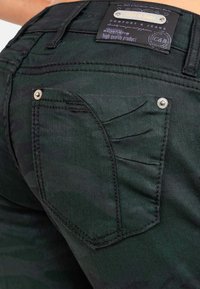 Primo piano di jeans in denim nero, che mostra una tasca posteriore con rivetti metallici e un'etichetta del marchio nera e argento cucita sulla vita.