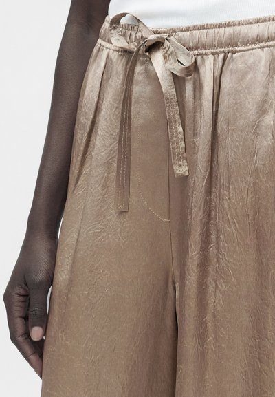 Κοντινή λήψη σκοτεινόχρωμου χεριού που ακουμπάει δίπλα σε άνετο, τσαλακωμένο μπεζ παντελόνι με δεμένη υφασμάτινη ζώνη στη ελαστική μέση.