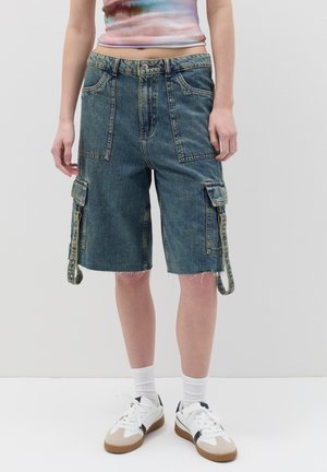 Denim cargo shorts in a faded blue wash, featuring multiple pockets, frayed hem, and adjustable side tabs. Paired with white sneakers.