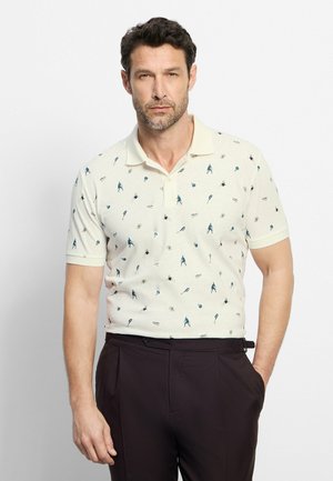 Uomo con polo bianca a maniche corte con piccole figure sportive blu e stampa di racchette, abbinata a pantaloni scuri, in piedi su sfondo bianco.