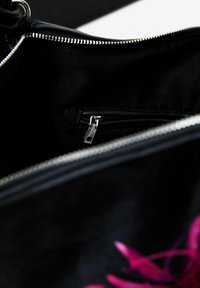 Czarna torebka od wewnątrz o gładkiej fakturze, z kieszenią na zamek błyskawiczny w kolorze srebrnym i różowymi kwiatowymi akcentami. Podszewka jest ciemna i minimalistyczna.