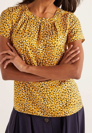 Kobieta nosząca żółtą bluzkę z krótkim rękawem w czarne i białe plamy, ze skrzyżowanymi ramionami, w połączeniu z ciemną spódnicą zapinaną na guziki.