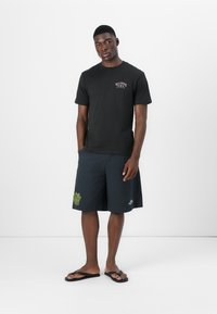 Zwarte katoenen t-shirt met een klein kleurrijk logo op de borst, gecombineerd met donkere shorts voorzien van groene bladborduursel. Zwarte slippers.