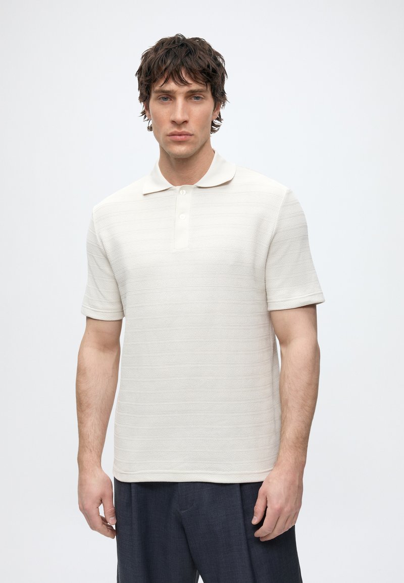 Jeune homme aux cheveux courts et bouclés portant un polo texturé blanc cassé à manches courtes et un pantalon foncé, debout devant un fond uni.