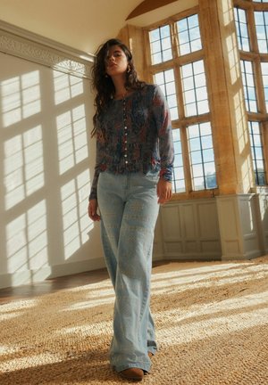 Frau mit langem Haar, die eine transparente Bluse mit Blumenmuster und weit geschnittene Jeans mit Muster trägt, steht in einem sonnenbeschienenen Zimmer mit großen Fenstern mit mehreren Scheiben.